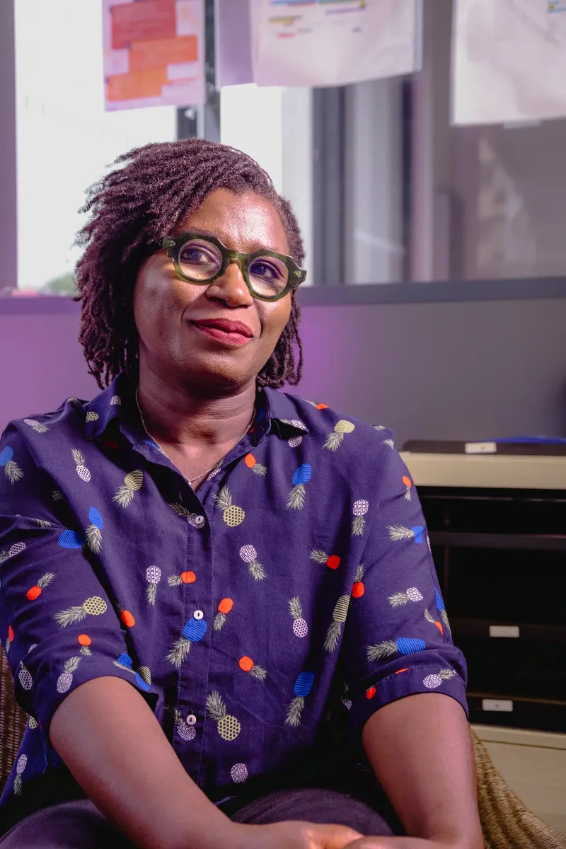 Portrait d’une femme assise dans son bureau, chemise à motifs, lunettes vertes, sourire léger et regard hors cadre